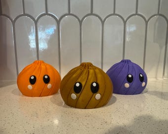 Puede incluir: Tres figuras redondeadas y en espiral en colores naranja, bronce y morado. Cada figura presenta una forma lisa de cúpula con un punto central superior, grandes ojos negros y pequeñas mejillas blancas circulares, creando un aspecto amigable.