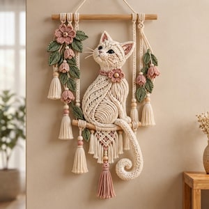 May include: A macrame wall hanging featuring a cream-colored cat sitting on a wooden dowel. The cat is adorned with a pink flower collar. The piece includes hanging vines with pink flowers, green leaves, and cream-colored tassels with wooden beads.