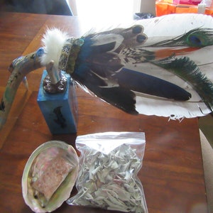 May include: A white feather fan with blue, green, and orange accents, a small wooden base, a white abalone shell with a pink stone inside, and a bag of white sage.