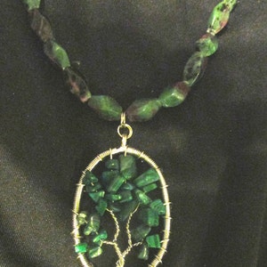 May include: A green gemstone necklace with a wire-wrapped tree pendant. The pendant is made of small green gemstones and wire, and is shaped like a tree with branches and leaves. The necklace is made of green gemstone beads.