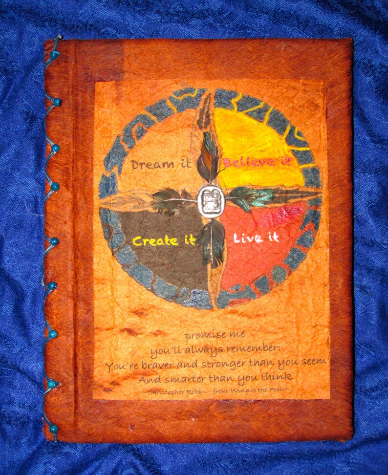 May include: A brown leather-bound journal with a colorful circular design on the front cover. The design features four sections with the words "Dream it", "Believe it", "Create it", and "Live it" written in white. The journal has a quote from Winnie the Pooh: "Promise me you'll always remember: You're braver and stronger than you seem and smarter than you think."