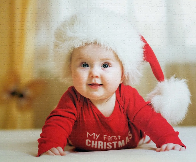 May include: A baby wearing a red and white Santa hat and a red long-sleeved onesie with the text "My First Christmas".