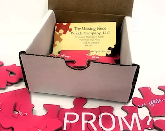 Può includere: Un puzzle a incastro rosa con la parola "PROM" in lettere bianche. I pezzi del puzzle sono sparsi intorno a una scatola bianca con un biglietto da visita all'interno. Il biglietto da visita dice "The Missing Piece Puzzle Company, LLC".