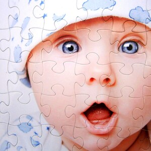 May include: A jigsaw puzzle featuring a close-up of a baby's face. The baby has wide blue eyes, a surprised expression, and is wearing a white hat with blue cloud patterns. The puzzle pieces are interlocked, forming the image.