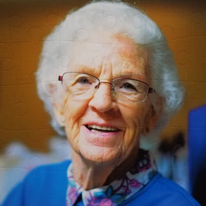 May include: A close-up portrait of a smiling older woman with white hair wearing glasses and a blue shirt with a floral pattern.