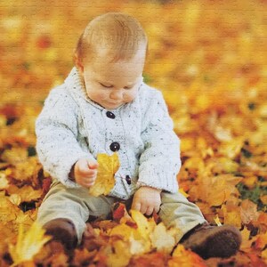 May include: A young child wearing a white sweater sits in a pile of autumn leaves. The child is holding a yellow leaf in their hand.