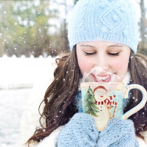 May include: A woman wearing a blue knitted hat and gloves is holding a blue and white ceramic mug with a snowman design. The mug is filled with a steaming beverage. The woman is standing in a snowy outdoor setting.