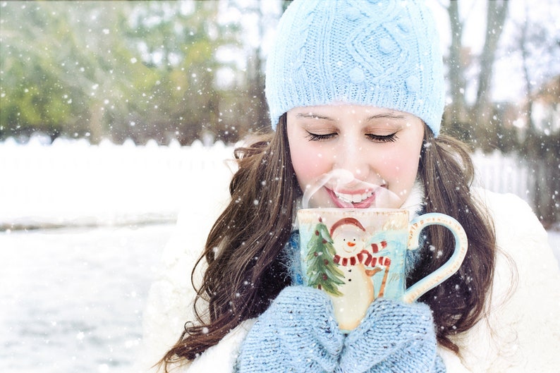 May include: A woman wearing a blue knitted hat and gloves is holding a blue and white ceramic mug with a snowman design. The mug is filled with a steaming beverage. The woman is standing in a snowy outdoor setting.