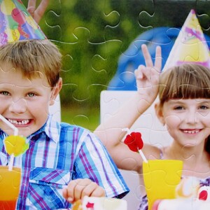 May include: A jigsaw puzzle featuring two children at a birthday party. The boy wears a blue plaid shirt and a party hat, holding a drink. The girl wears a party hat and makes a peace sign. Drinks and a cake are visible.