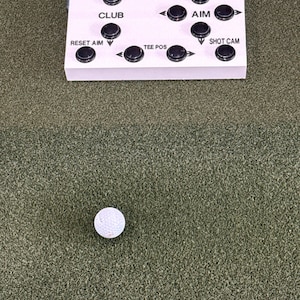 May include: A white rectangular control box with black circular buttons and printed labels including MULLIGAN, HEAT MAP, PUTT, FLYOVER, CLUB, AIM, RESET AIM, TEE POS, and SHOT CAM, positioned above a single white golf ball on green turf.