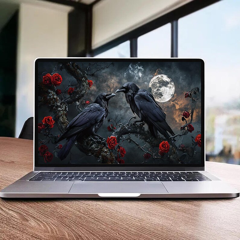 May include: A silver laptop displays a digital art scene of two black ravens perched on a tree branch with red roses. A full moon glows in the background, set against a dark, cloudy sky. The laptop sits on a wooden table.