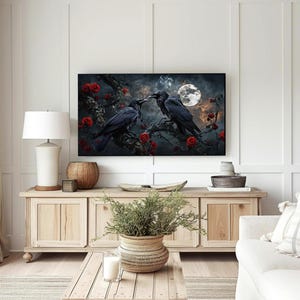 May include: A framed artwork depicts two black crows perched on a branch with red roses, under a full moon. The artwork is displayed above a wooden cabinet in a living room setting, with a white lamp and decorative items.