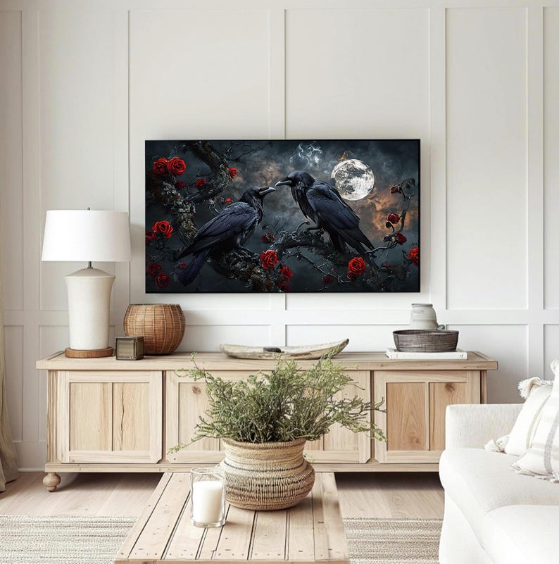 May include: A framed artwork depicts two black crows perched on a branch with red roses, under a full moon. The artwork is displayed above a wooden cabinet in a living room setting, with a white lamp and decorative items.