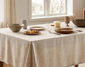 May include: A beige textured tablecloth covers a dining table set with a light gray speckled mug, a tan bowl, a small plate with food, a brown plate, a square candle, and a dark brown bowl. A silver fork rests on the table.