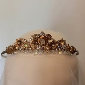May include: A gold tiara with a floral design and clear rhinestones. The tiara is on a white mannequin head.
