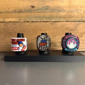 May include: Three championship rings displayed on black stands. The left ring features Big League Chew branding and a baseball player. The center ring has a silver eagle emblem with crystals. The right ring is pink and blue with Tournament Champions text.