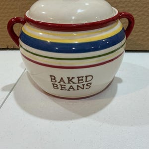 May include: A cream-colored ceramic bean pot with a matching lid and two red side handles. The pot features horizontal stripes in yellow, blue, and green, with the words BAKED BEANS printed in brown lettering on the front surface.