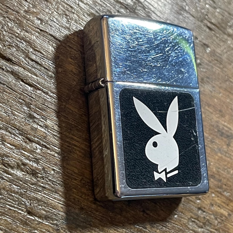 K&ouml;nnte beinhalten: Ein silberfarbenes Metallfeuerzeug mit einem schwarzen rechteckigen Emblem mit dem wei&szlig;en Playboy-Hasenlogo. Das Feuerzeug hat eine rechteckige Form und einen Klappdeckel. Der Hintergrund ist eine Holzoberfl&auml;che.