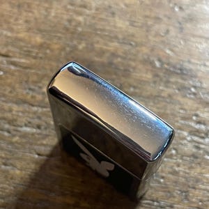 K&ouml;nnte beinhalten: Ein silberfarbenes Feuerzeug mit einem schwarzen Feld, das ein wei&szlig;es Hasenlogo zeigt. Das Feuerzeug hat eine gl&auml;nzende, reflektierende Oberfl&auml;che und eine rechteckige Form. Das Bild ist leicht schr&auml;g aufgenommen und zeigt die Ober- und Seite des Feuerzeugs.