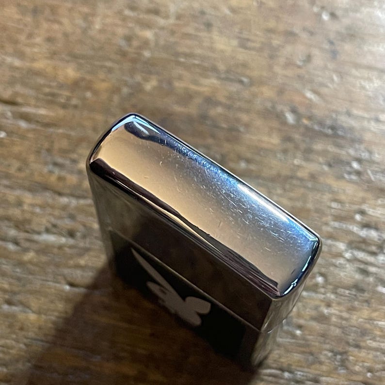 K&ouml;nnte beinhalten: Ein silberfarbenes Feuerzeug mit einem schwarzen Feld, das ein wei&szlig;es Hasenlogo zeigt. Das Feuerzeug hat eine gl&auml;nzende, reflektierende Oberfl&auml;che und eine rechteckige Form. Das Bild ist leicht schr&auml;g aufgenommen und zeigt die Ober- und Seite des Feuerzeugs.