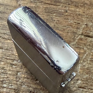 May include: A shiny, chrome-colored lighter with a rectangular shape. The lighter has a hinged lid and a small, metallic lever on the side. The surface shows minor wear and tear, with small imperfections. The lighter is set against a wooden background.