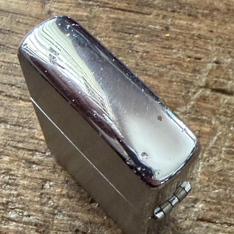 May include: A shiny, chrome-colored lighter with a rectangular shape. The lighter has a hinged lid and a small, metallic lever on the side. The surface shows minor wear and tear, with small imperfections. The lighter is set against a wooden background.