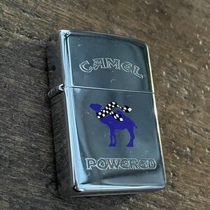 May include: A silver-colored lighter with the word "CAMEL" arched across the top. Below is a purple camel wearing a black and white checkered scarf. The word "POWERED" is printed below the camel.