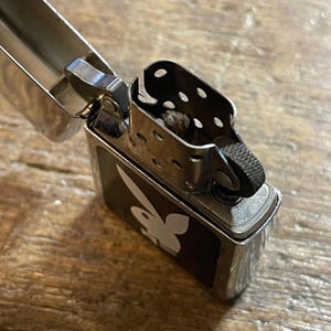 K&ouml;nnte beinhalten: Ein silbernes Metallfeuerzeug mit offenem Deckel, das das Innere freilegt. Das Feuerzeug hat eine schwarze rechteckige Platte mit einem wei&szlig;en Playboy-Hasenlogo. Das Feuerzeug liegt auf einer Holzoberfl&auml;che.
