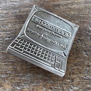 May include: A silver-colored computer-shaped pin with the text "AG COMMAND Computer System SPERRY NEW HOLLAND" is displayed. The pin has a keyboard design at the bottom and a screen at the top. The pin is on a wooden surface.