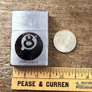May include: A silver-colored lighter with a black and white 8-ball design. The lighter has the text "&copy; 1993 A.A.D.L.P." A quarter and a ruler are next to the lighter.
