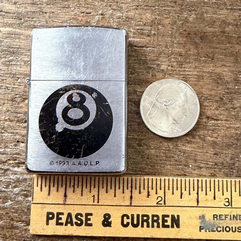 May include: A silver-colored lighter with a black and white 8-ball design. The lighter has the text "&copy; 1993 A.A.D.L.P." A quarter and a ruler are next to the lighter.