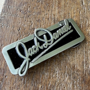 May include: A rectangular belt buckle with a brushed silver-tone metal frame and a black enamel inlay. The raised silver-tone script text reads Jack Daniel across the center of the buckle, featuring a smooth finish and a classic rectangular shape.