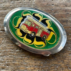 May include: Oval silver-tone belt buckle with a red and yellow fire truck design. The fire truck is set against a green background with yellow swirls. The buckle is a vintage accessory.