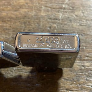 K&ouml;nnte beinhalten: Nahaufnahme eines gl&auml;nzenden, silbernen Zippo-Feuerzeugs. Auf dem Feuerzeug sind das Zippo-Logo und der Text "BRADFORD, PA. MADE IN U.S.A." eingraviert. Das Feuerzeug liegt auf einer Holzoberfl&auml;che.