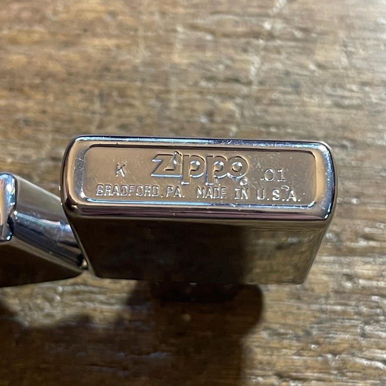 K&ouml;nnte beinhalten: Nahaufnahme eines gl&auml;nzenden, silbernen Zippo-Feuerzeugs. Auf dem Feuerzeug sind das Zippo-Logo und der Text "BRADFORD, PA. MADE IN U.S.A." eingraviert. Das Feuerzeug liegt auf einer Holzoberfl&auml;che.