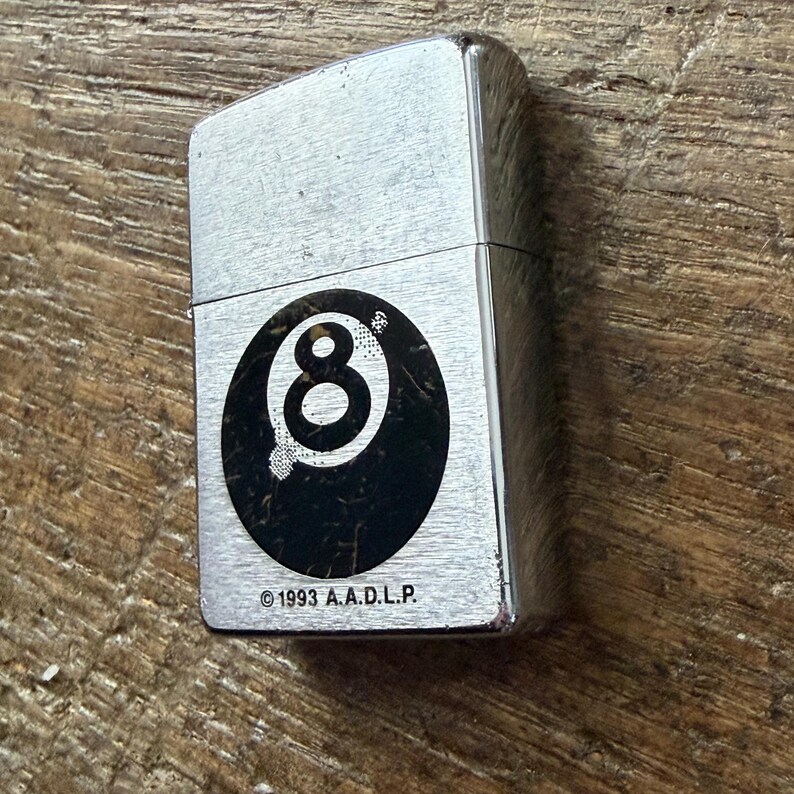 May include: A silver-colored lighter with a black and white eight-ball graphic. The lighter has a rectangular shape and a hinged lid. The text "&copy;1993 A.A.D.L.P." is printed below the graphic.