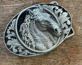 May include: Silver-toned metal belt buckle featuring a detailed horse head within an oval frame. The horse head is textured, and the frame has a black background with ornate scrollwork. A classic Western-style accessory.
