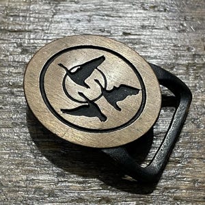 May include: A brass-colored belt buckle with a black frame. The buckle features a circular design with three stylized birds in flight. The birds are black against a lighter background, creating a contrasting visual.