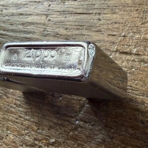 May include: A silver Zippo lighter with the brand name and location of manufacture, Bradford, PA, Made in U.S.A., engraved on the bottom. The lighter is rectangular with rounded edges and a brushed metal finish.