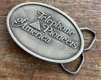 May include: Oval-shaped silver-toned belt buckle with the words "Telephone Pioneers of America" in raised lettering. The buckle has a textured surface and a loop for the belt. A triangle with a design is between the words.