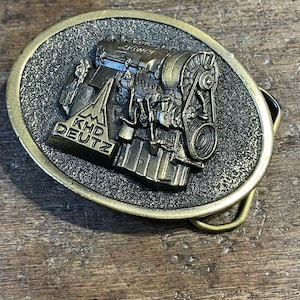 May include: Oval-shaped, antique brass belt buckle featuring a detailed relief of an engine. The buckle has a textured background and the text "KHD DEUTZ" is visible on the engine's structure. The buckle is designed for a belt.