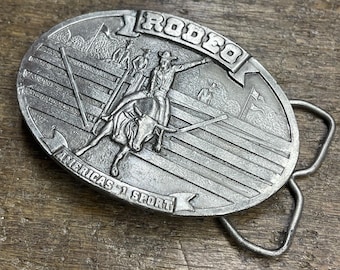 May include: Oval-shaped silver belt buckle with a rodeo theme. The buckle features a cowboy riding a bull, with the word "RODEO" at the top and "AMERICA'S 1 SPORT" at the bottom. The buckle has a silver finish.