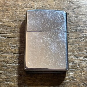 K&ouml;nnte beinhalten: Ein silberfarbenes, rechteckiges Feuerzeug mit geb&uuml;rsteter Metalloberfl&auml;che. Das Feuerzeug hat einen Klappdeckel und ein kleines Metallscharnier an der Seite. Die Oberfl&auml;che weist leichte Kratzer und Abnutzungserscheinungen auf.