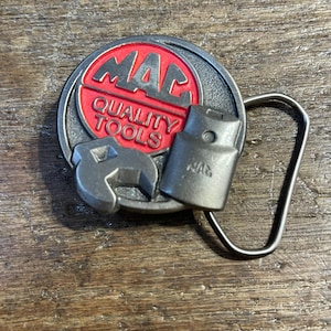 May include: A silver-colored metal belt buckle with the words "MAC QUALITY TOOLS" in red and white. The buckle features a wrench and a tool, and the brand name "MAC" is visible.