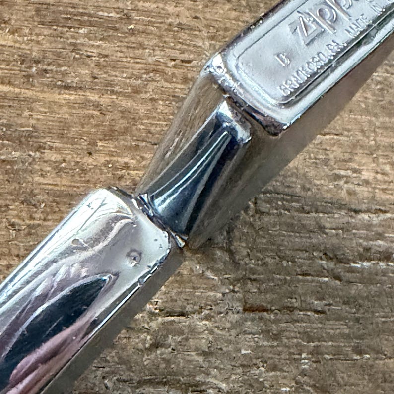 May include: Close-up of a chrome-colored Zippo lighter. The lighter is highly reflective, with visible text that reads "Zippo" and "BRADFORD, PA. MADE". The lighter is on a wooden surface.