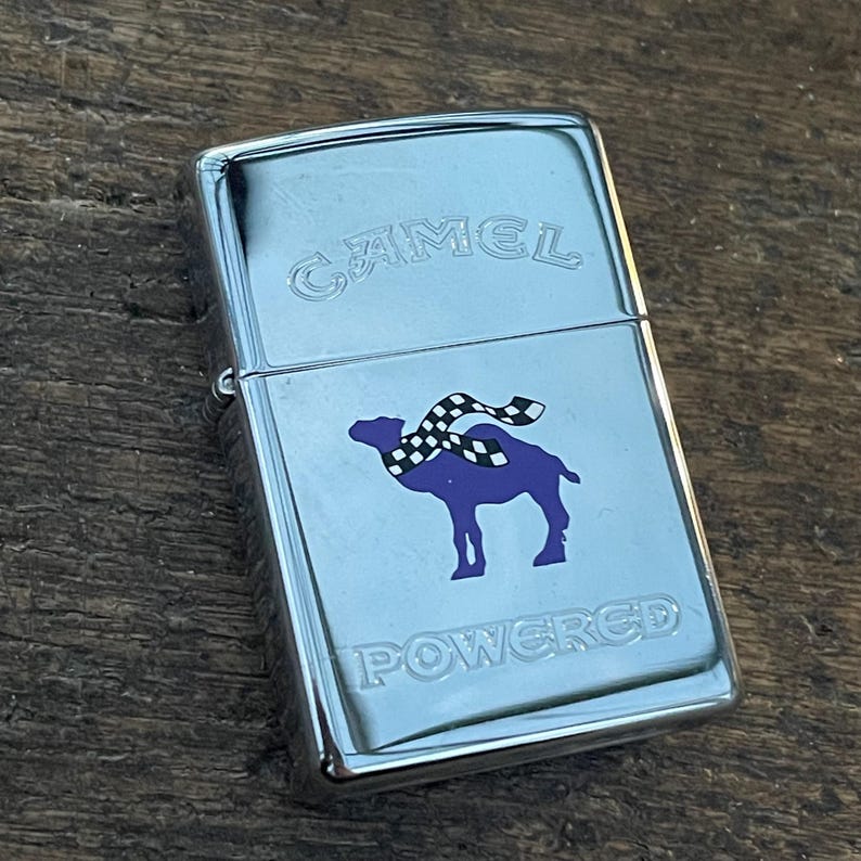 May include: A chrome-plated lighter featuring the Camel logo. The design includes the word "CAMEL" at the top and a purple camel with a checkered scarf. The word "POWERED" is printed below the camel.