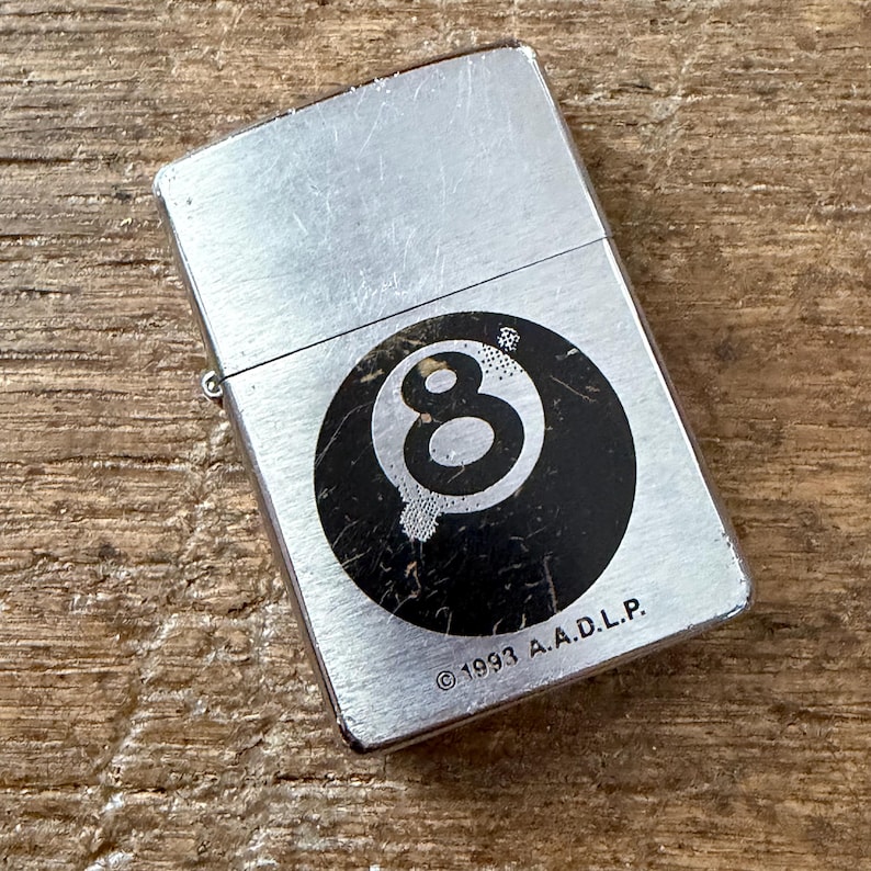 May include: A silver-toned metal lighter with a black and white eight ball graphic. The lighter has a rectangular shape and a hinged lid. The text "&copy;1993 A.A.D.L.P." is printed below the graphic.