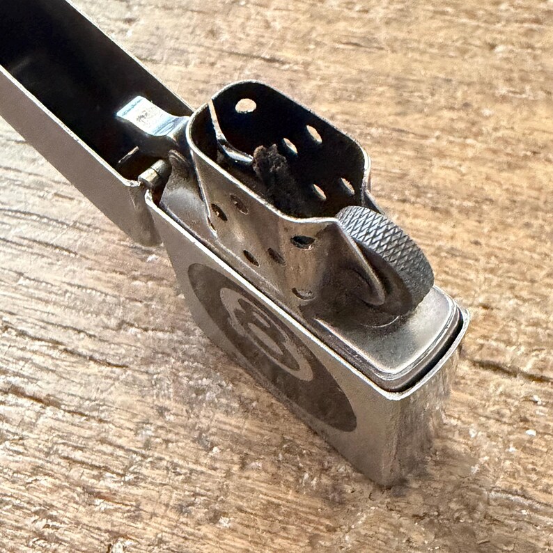 May include: A silver-colored metal lighter with an open lid, revealing the wick and flint wheel. The lighter has a circular design on the front. The lighter is on a wooden surface.