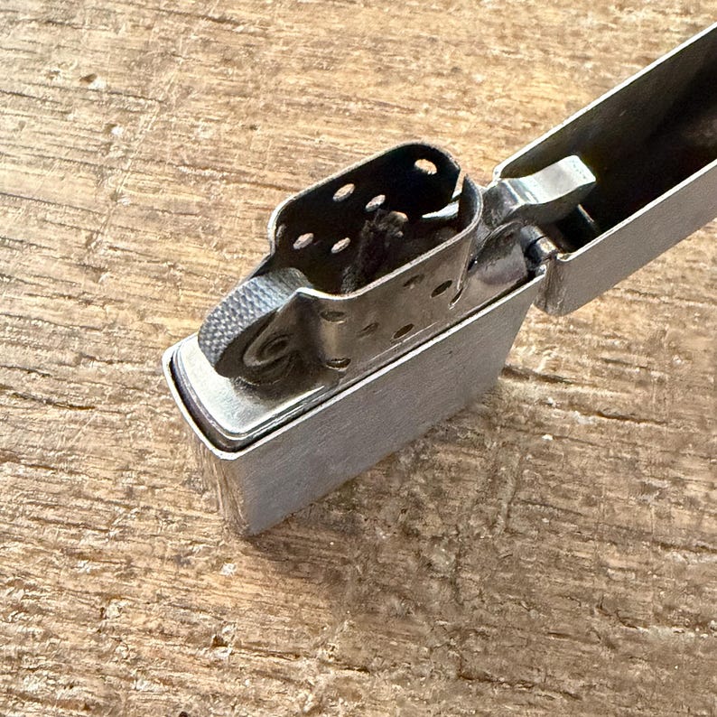 May include: A silver metal lighter with the top open, revealing the internal components. The lighter has a textured wheel for sparking and a hinged lid. The background is a wooden surface.