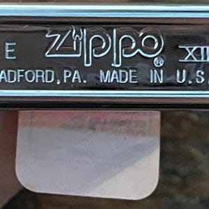 May include: A chrome-finished Zippo lighter base with the brand name and "BRADFORD, PA. MADE IN U.S.A." printed in white. The lighter has a rectangular shape with rounded edges. The Roman numeral XIII is also visible.